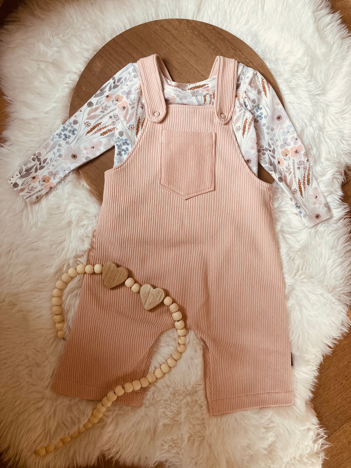 Een baby's Lichte perzikkleurige ribstof overall en een ketting aan een kleedje van Cuteez.